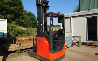 A bright orange forklift is parked on a sunny day next to a building and stacks of wooden planks. Green foliage and open sky are visible in the background.