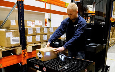 A warehouse worker places a package marked with a hazard symbol onto a scanning platform amid shelves of labeled boxes.