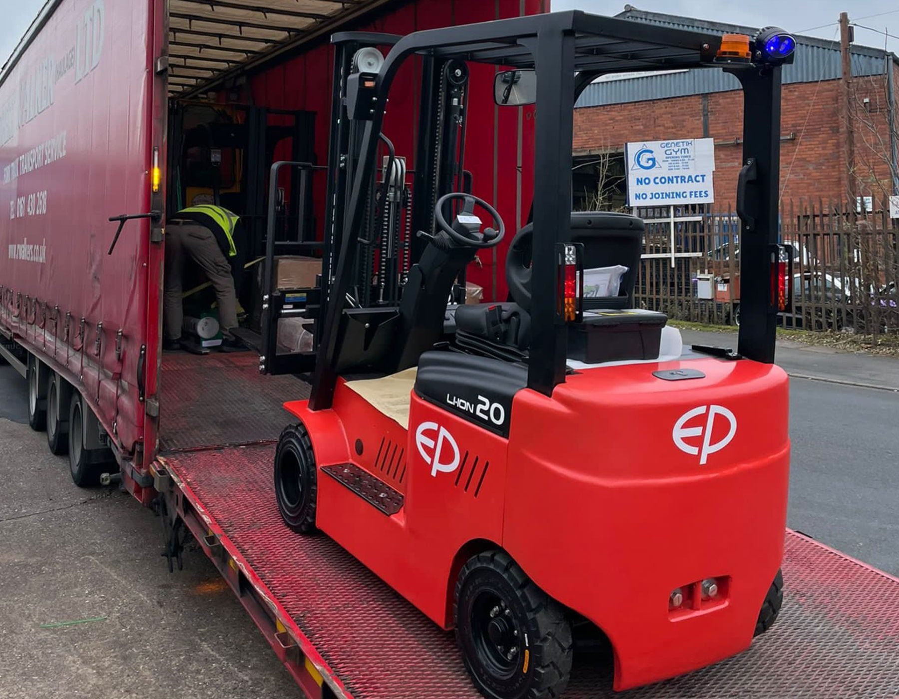 Red EP electric forklift parked on a loading ramp next to an open truck, with a worker in a high-visibility vest preparing items for transport, in an outdoor industrial setting.