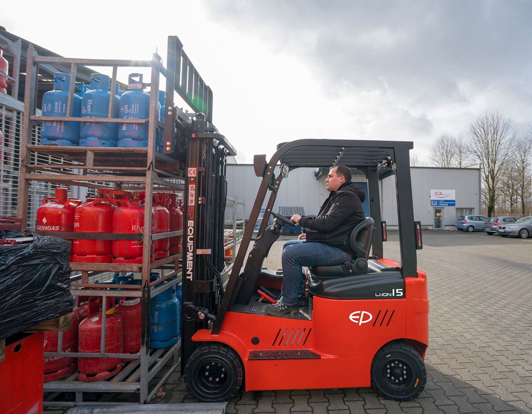 Operator driving a red EP electric forklift, transporting a rack loaded with blue and red gas cylinders in an outdoor industrial area, with buildings and a cloudy sky in the background.