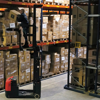 Warehouse worker using a red electric order picker to lift and stack boxes on high shelves, surrounded by rows of palletized goods and marked storage sections.