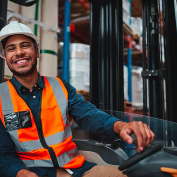 Smiling warehouse worker in a high-visibility orange vest and white hard hat, sitting in a forklift inside a warehouse, with shelving and equipment in the background.