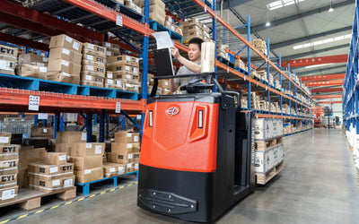 A worker drives a red order picker forklift in a warehouse, checking a paper list while surrounded by shelves filled with boxes.