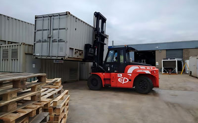 A red forklift is lifting a large shipping container in an industrial yard. Wooden pallets are stacked nearby, under a cloudy sky. Depicts heavy machinery in action.