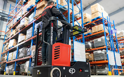 A worker operates a red electric pallet truck in a busy warehouse, surrounded by high shelves filled with boxes and pallets.