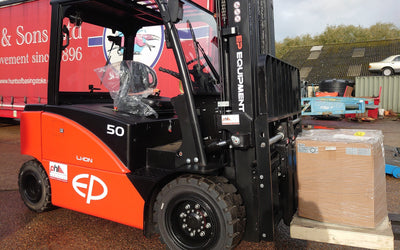 A red and black EP Equipment forklift lifts a large cardboard box on a pallet outdoors on a wet surface, with a red truck trailer in the background.