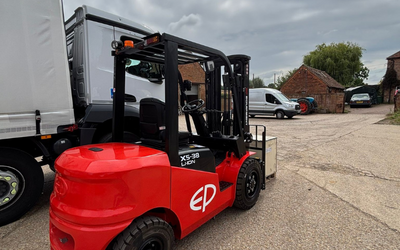 Red EP forklift parked near a white truck on a gravel lot under a cloudy sky with buildings and vehicles in the background.