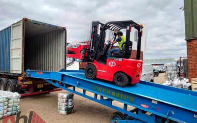 A forklift is loading goods onto a blue ramp from a shipping container, surrounded by stacks of bags on the ground.