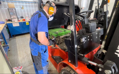 A technician in blue coveralls repairs the engine of a forklift, wearing safety headphones and a mask in a workshop environment.