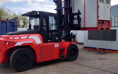Red and black forklift lifting a large white and red industrial container in an outdoor storage area under a partly cloudy sky.