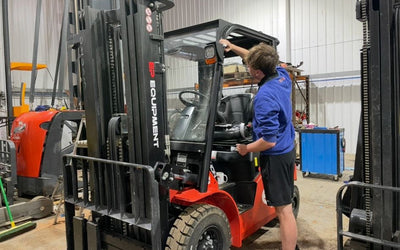 A worker in a blue sweatshirt inspects the cabin of a red forklift in a well-lit warehouse, surrounded by various equipment.