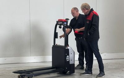 Two individuals stand near a black electric pallet jack in an empty warehouse, discussing its features against a plain white wall.