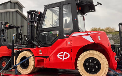 A red forklift with white tires is secured on a flatbed trailer in an outdoor industrial area under a cloudy sky.