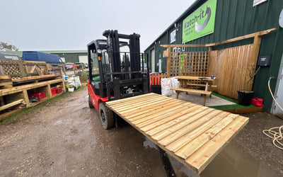 A red forklift carries a large wooden pallet in a muddy outdoor area. Surrounding structures include a green building, storage racks, and a picnic table.