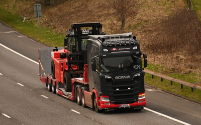 Black and red Scania truck carrying heavy red forklift on a flatbed trailer driving on a highway.