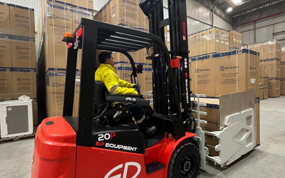 A forklift operator in a yellow jacket maneuvers a red EP Equipment forklift among tall stacks of cardboard boxes in a warehouse.