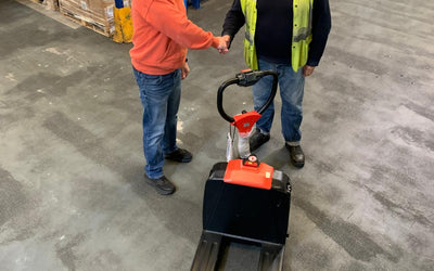 Two workers shake hands in a warehouse, with a pallet jack prominently displayed on the floor amidst stacked pallets in the background.