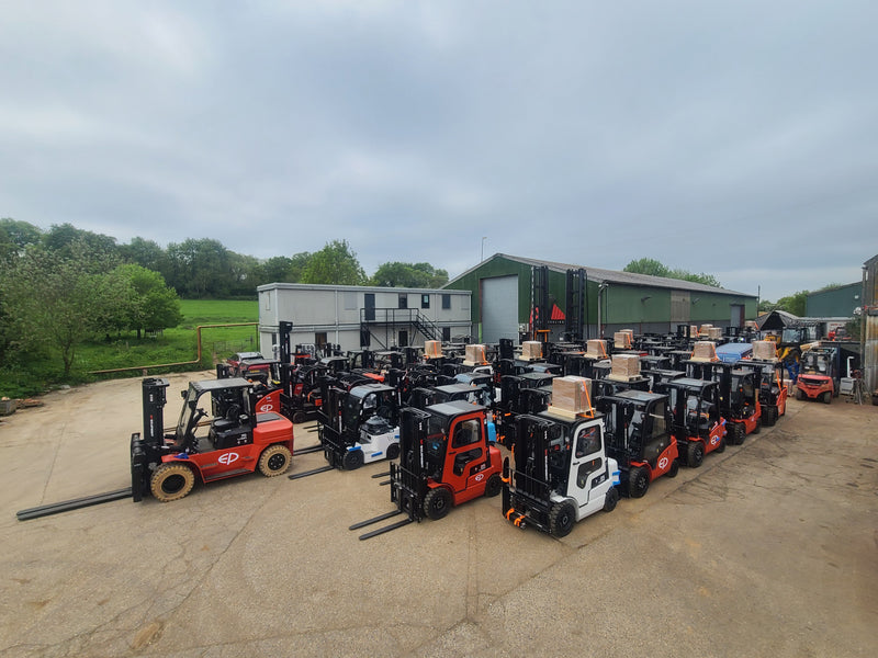 Row of forklifts with cargo on a concrete surface with a building and trees in the background.