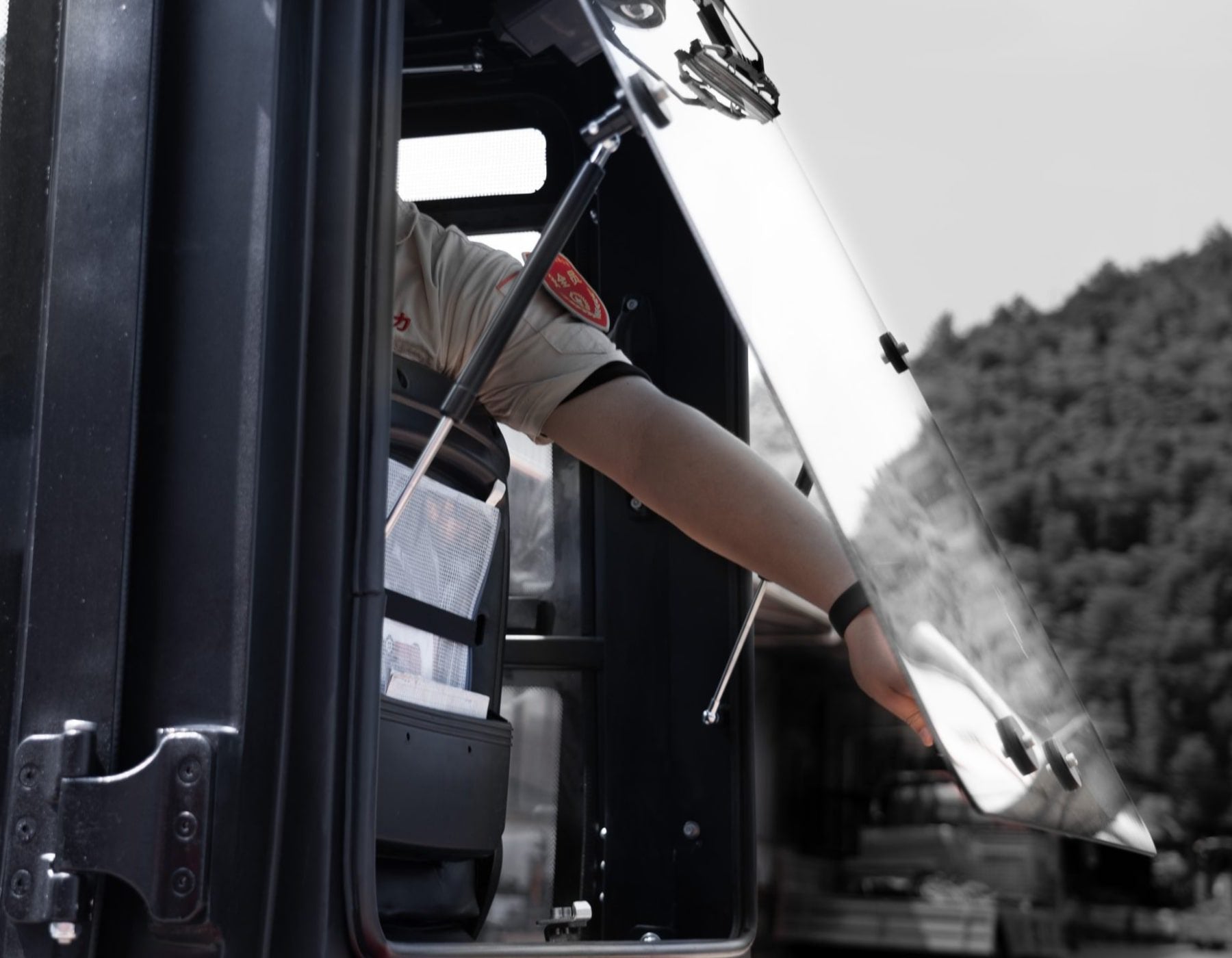 Close-up of a forklift operator's arm reaching out of the cab, holding onto an open protective canopy, with a blurred background of trees and outdoor surroundings.