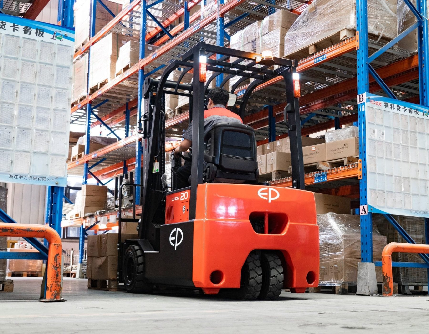 Rear view of a red and black EP electric forklift being operated in a warehouse aisle, maneuvering between tall blue shelves stacked with pallets and boxes, with signage and protective barriers in the background.