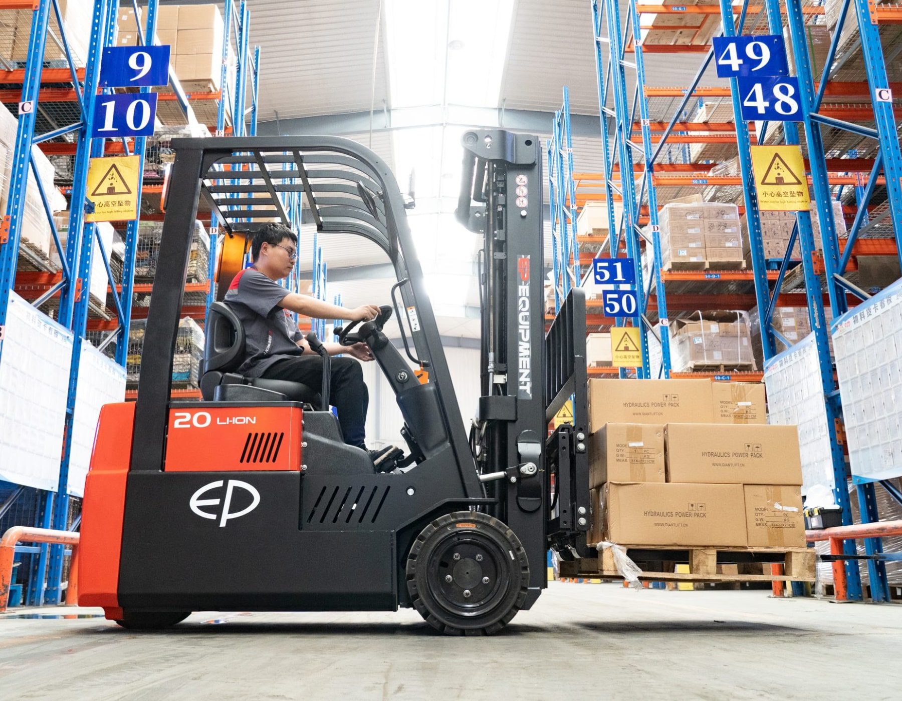 Warehouse worker driving a red and black EP electric forklift, transporting a pallet loaded with boxes through a spacious aisle marked with numbered blue signage, surrounded by tall shelves filled with inventory.