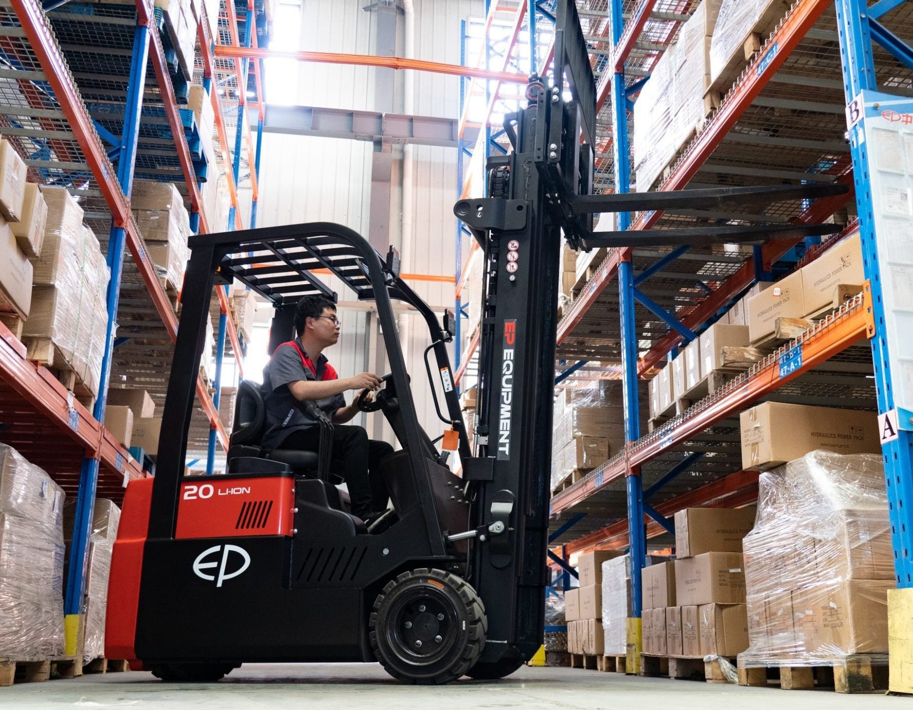 Side view of a warehouse worker operating a red and black EP electric forklift, lifting forks positioned to retrieve or place boxes on tall blue shelving units filled with stacked goods.