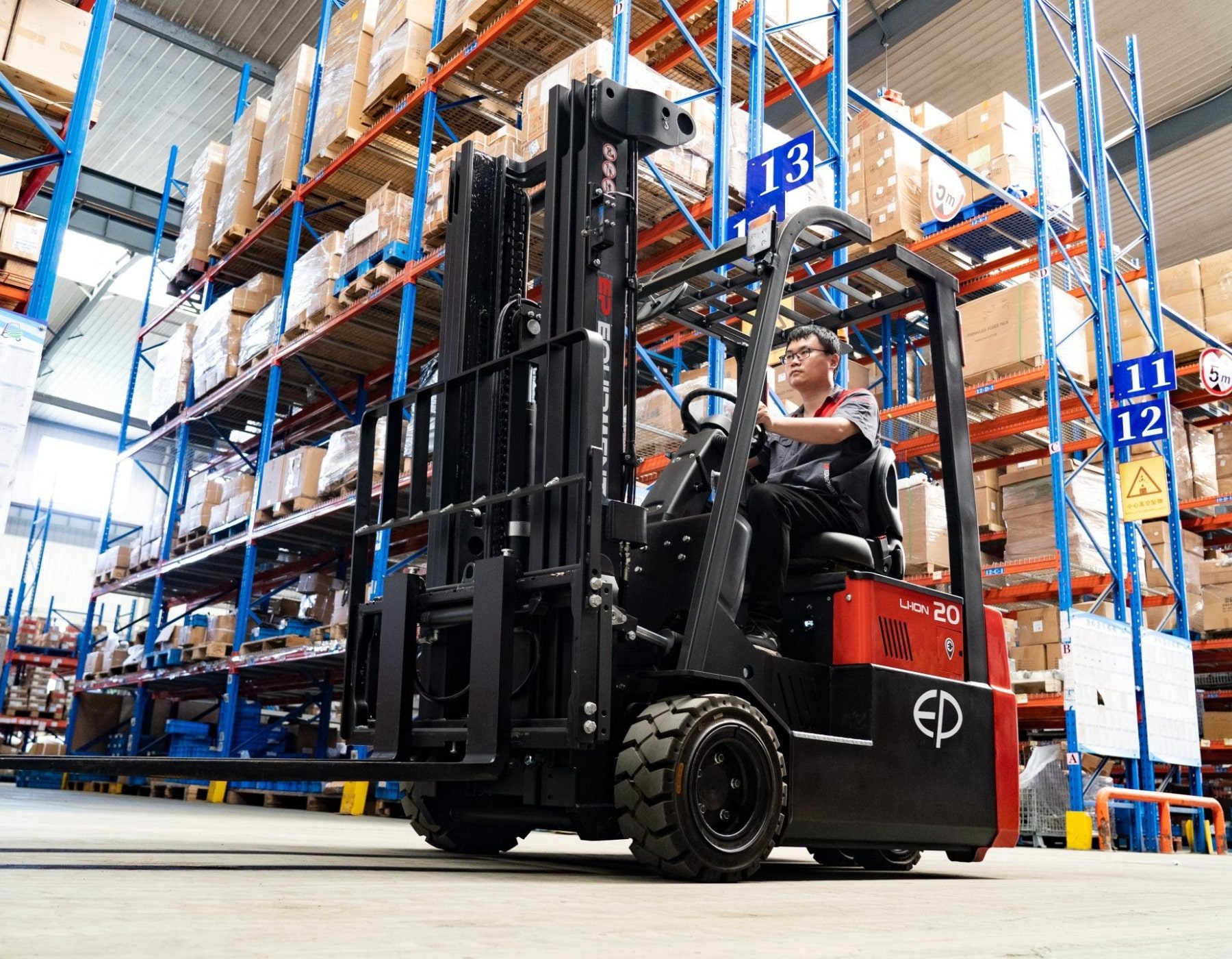 Warehouse operator driving a red and black EP electric forklift through a large aisle with blue shelving filled with stacked boxes, under a high ceiling and numbered signs indicating storage sections.