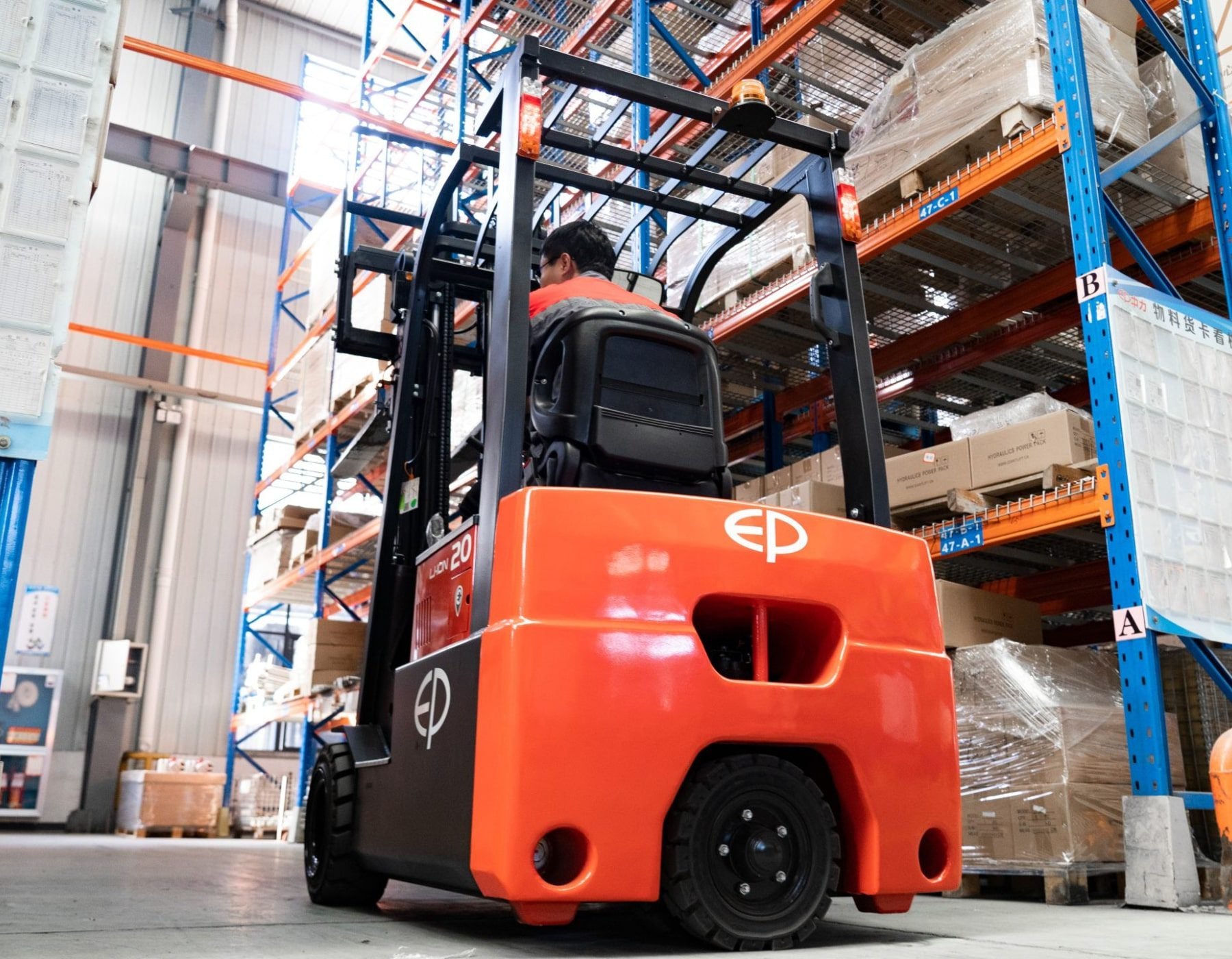 Rear view of a red and black EP electric forklift, with an operator navigating through a warehouse aisle lined with blue and orange shelving filled with palletized boxes.