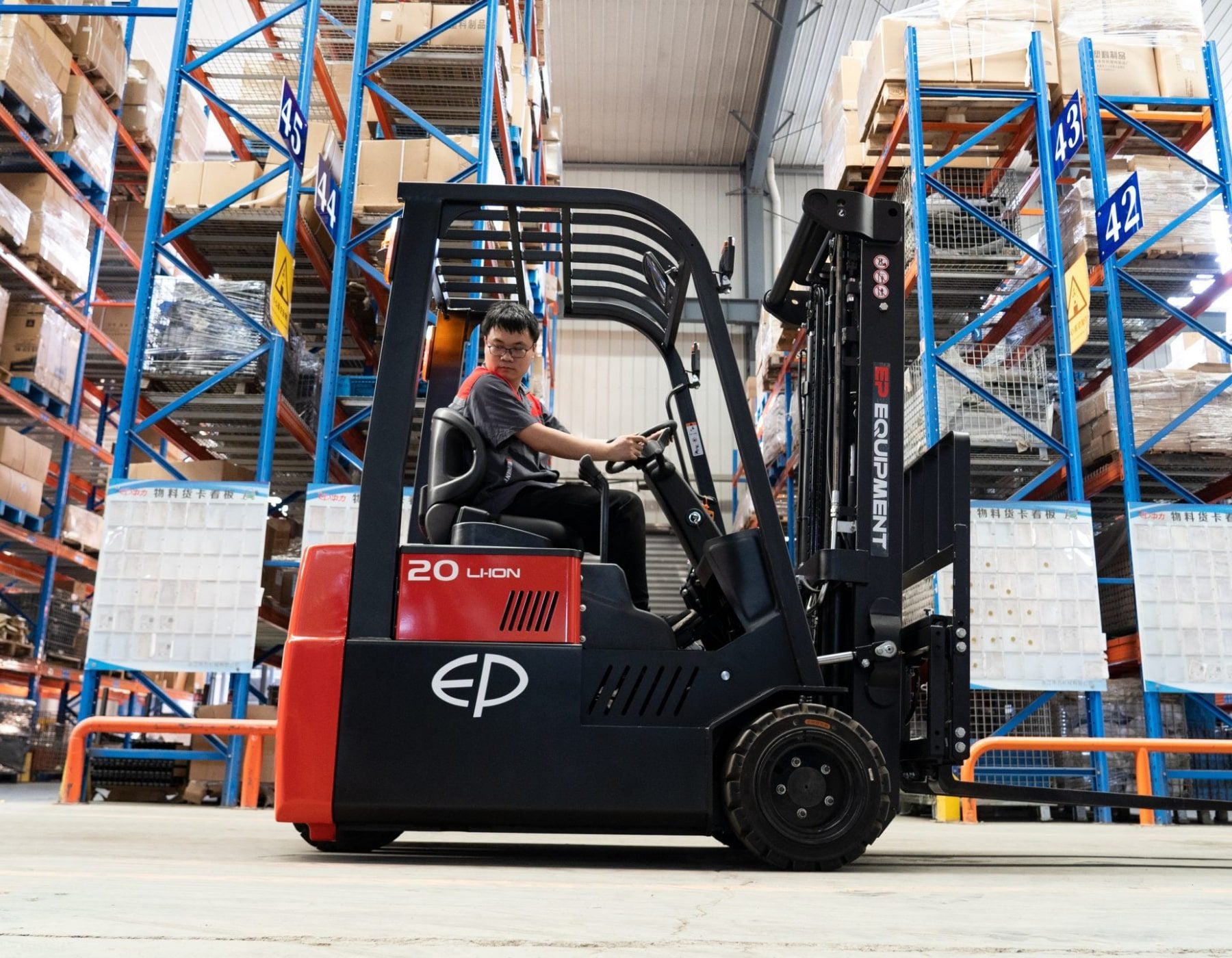 Side view of a warehouse worker operating a red and black EP electric forklift, driving through an aisle lined with tall blue shelving stocked with pallets and boxes, under a high industrial ceiling.