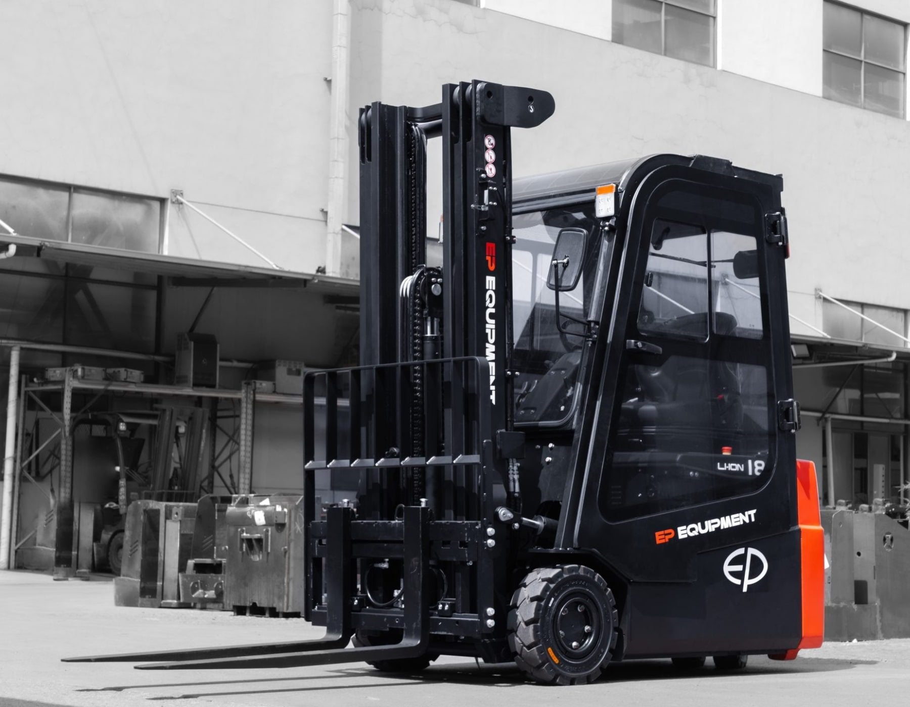 Black and red EP electric forklift parked outside an industrial building, featuring a protective enclosed cab and sturdy lifting forks, with equipment and structural elements in the background.