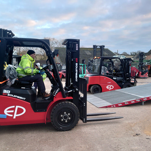 Red forklift with a person operating it in an outdoor setting with other forklifts and equipment.