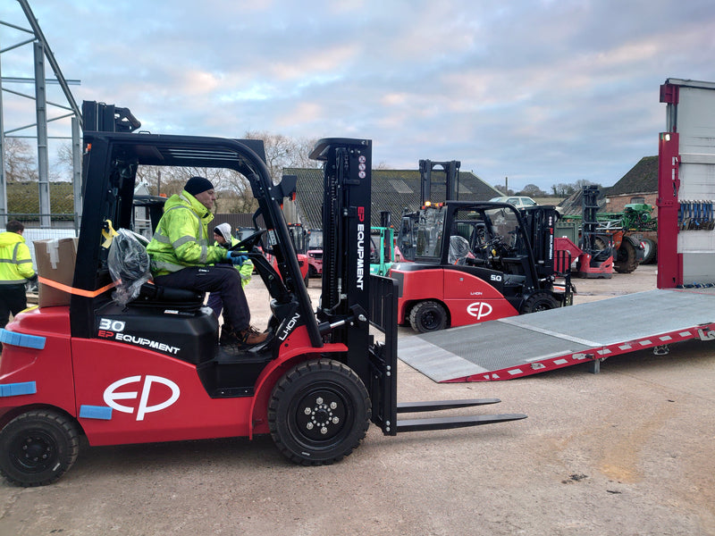 Red forklift with a person operating it in an outdoor setting with other forklifts and equipment.