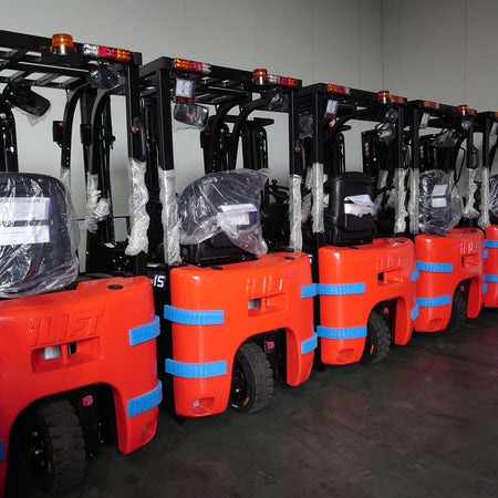 A row of new orange electric forklifts, covered in plastic and secured with blue straps, awaiting delivery in a warehouse.