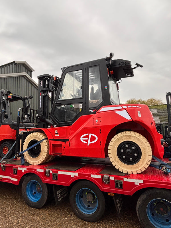 Red electric forklift with solid tires secured on a flatbed trailer, parked near industrial buildings under cloudy sky.