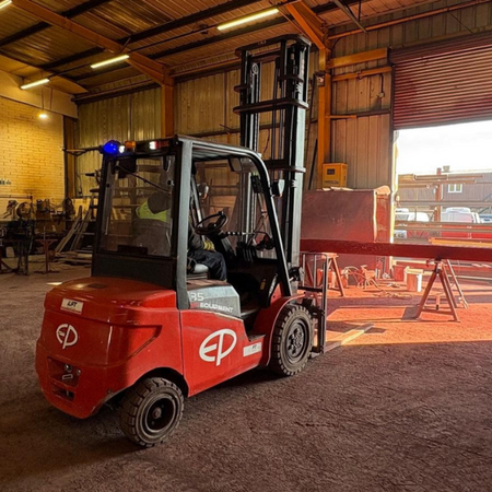 A red forklift is seen in a well-lit warehouse, maneuvering near metal workstations and a large opened door revealing daylight.
