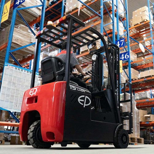 Worker operating a red and black forklift in a large warehouse with tall blue shelving units stocked with boxed and palletized goods. The warehouse aisles are marked with visible signage and numbers.