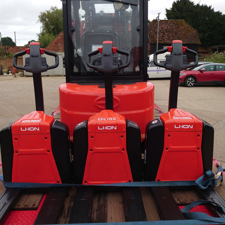 Three red electric pallet jacks with 'EPL185' labels secured on a truck bed, with a background of parked vehicles and greenery.